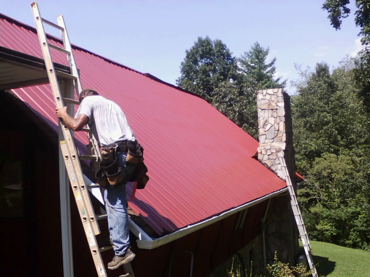 Roofer on a ladder beside a red metal roof and stone chimney — Soffit & Fascia Repair by Roofing Brothers in Pekin